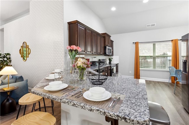 kitchen with light stone counters, appliances with stainless steel finishes, a kitchen breakfast bar, lofted ceiling, and light hardwood / wood-style flooring