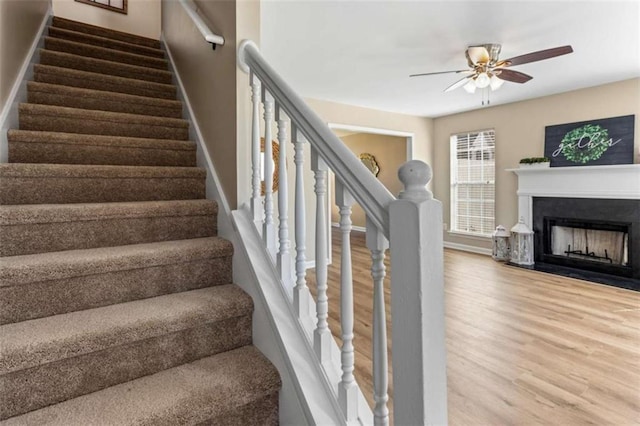 stairway featuring a fireplace with flush hearth, a ceiling fan, baseboards, and wood finished floors