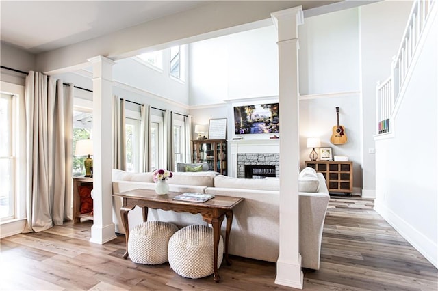 living area with wood finished floors, a fireplace, ornate columns, baseboards, and a high ceiling
