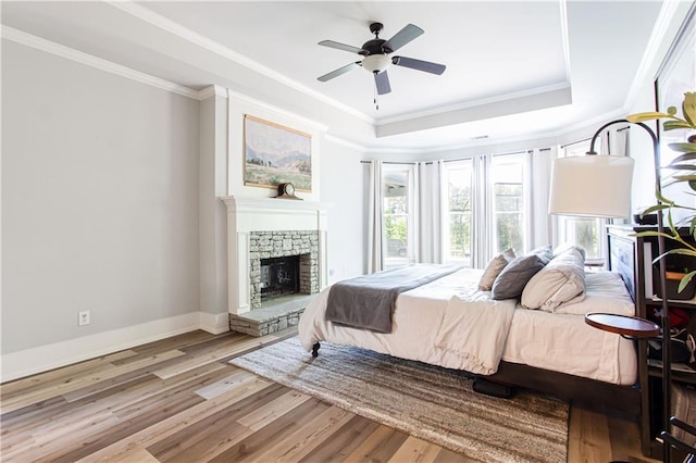 bedroom with a raised ceiling, wood finished floors, crown molding, baseboards, and a stone fireplace