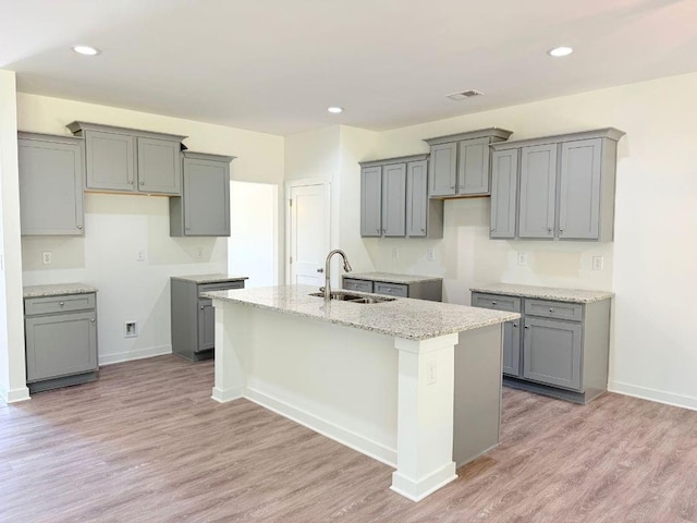 kitchen featuring an island with sink, light stone counters, gray cabinets, a sink, and light wood-style floors