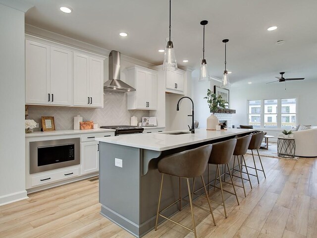 kitchen with built in microwave, white cabinetry, a kitchen island with sink, sink, and wall chimney exhaust hood