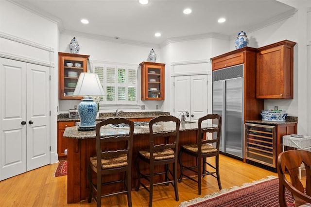 kitchen featuring stainless steel built in fridge, wine cooler, ornamental molding, an island with sink, and light hardwood / wood-style floors