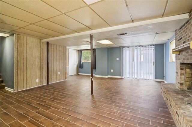 basement with a brick fireplace, wooden walls, a paneled ceiling, and dark hardwood / wood-style flooring