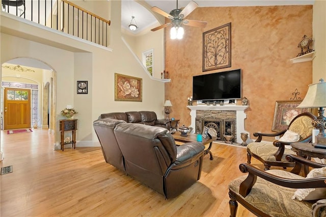 living area featuring light wood finished floors, arched walkways, a towering ceiling, ceiling fan, and a stone fireplace