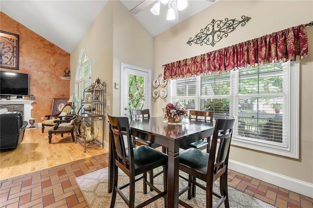 dining area with baseboards, ceiling fan, brick floor, a fireplace, and high vaulted ceiling