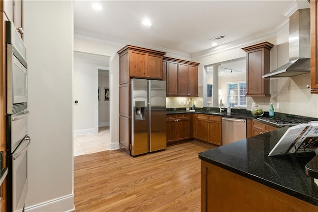 kitchen with appliances with stainless steel finishes, wall chimney range hood, dark stone counters, kitchen peninsula, and light wood-type flooring