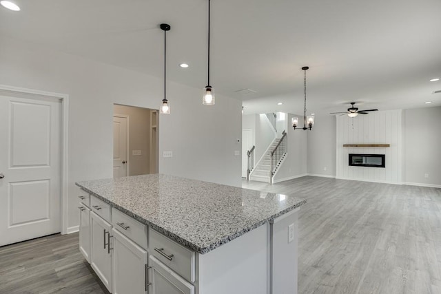 kitchen with light stone counters, a large fireplace, ceiling fan with notable chandelier, white cabinetry, and light wood finished floors