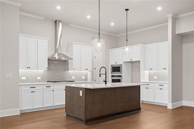 kitchen featuring wall chimney range hood, a kitchen island with sink, stainless steel appliances, white cabinets, and decorative light fixtures