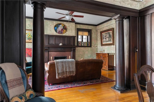 living area featuring ceiling fan, light wood-style flooring, wallpapered walls, decorative columns, and crown molding