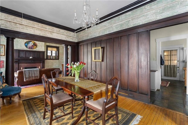 dining area with light wood-style flooring, a notable chandelier, ornamental molding, ornate columns, and wallpapered walls