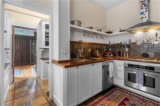 kitchen with stainless steel appliances, a sink, wood counters, white cabinetry, and wall chimney range hood