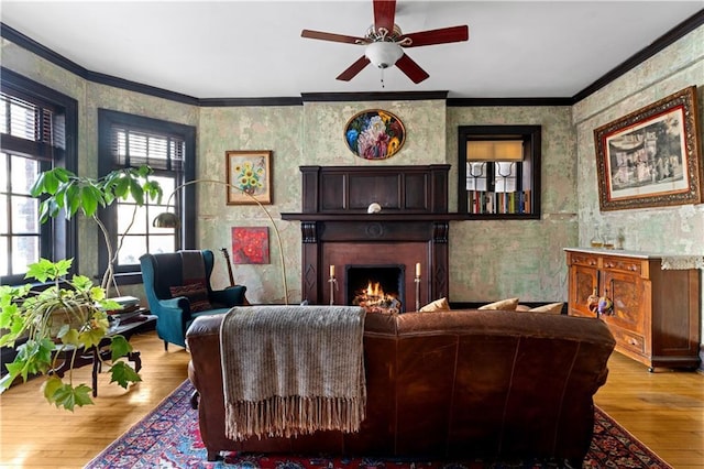 living area with a warm lit fireplace, wallpapered walls, a ceiling fan, light wood-style flooring, and ornamental molding