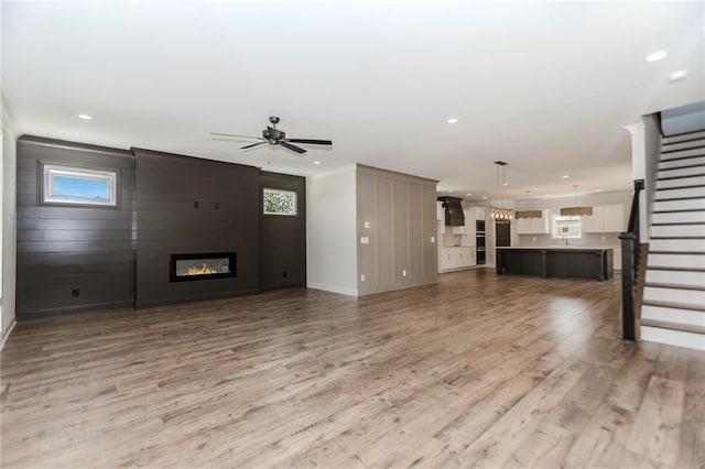 unfurnished living room with stairs, light wood-style floors, baseboards, a large fireplace, and a ceiling fan