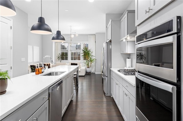 kitchen with appliances with stainless steel finishes, wall chimney exhaust hood, sink, gray cabinets, and hanging light fixtures