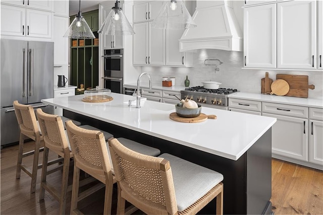 kitchen with appliances with stainless steel finishes, an island with sink, a kitchen breakfast bar, hanging light fixtures, and custom range hood