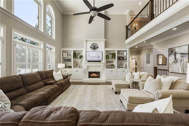 living room with a towering ceiling, ceiling fan, ornamental molding, wood finished floors, and a lit fireplace