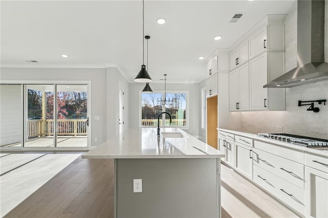 kitchen with wall chimney exhaust hood, hanging light fixtures, light hardwood / wood-style flooring, an island with sink, and white cabinets