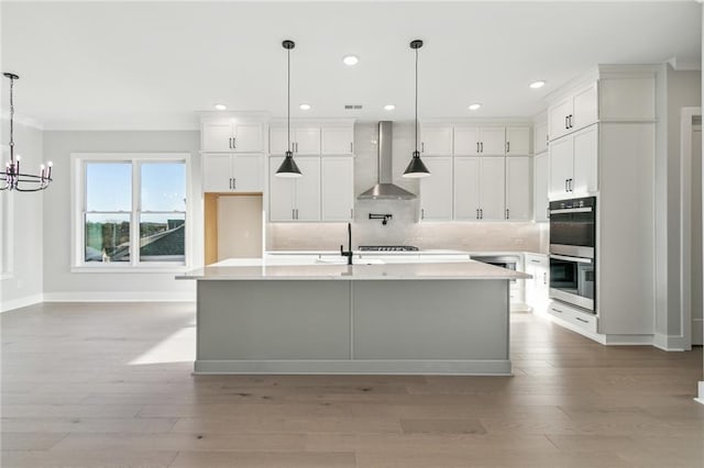 kitchen featuring wall chimney exhaust hood, stainless steel double oven, white cabinets, hanging light fixtures, and an island with sink