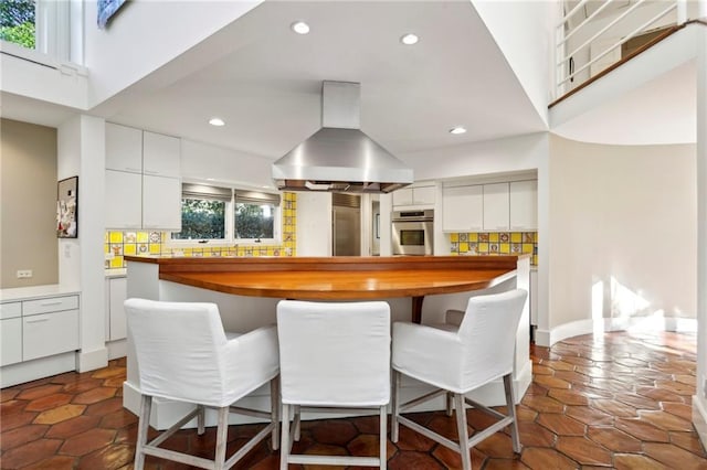 kitchen with white cabinetry, stainless steel oven, backsplash, island exhaust hood, and a breakfast bar area