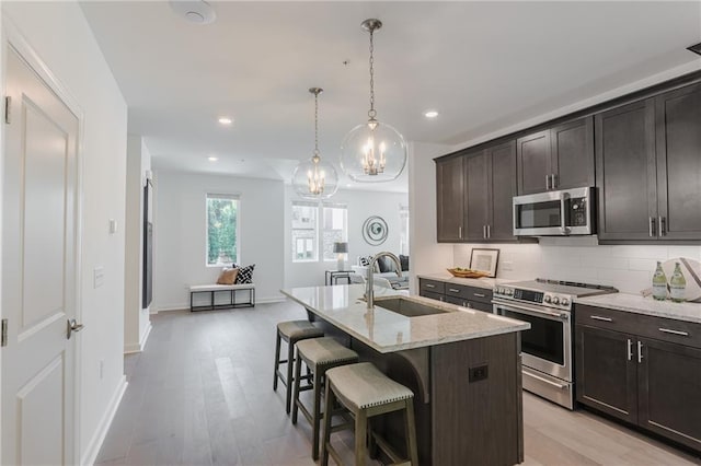 kitchen with sink, appliances with stainless steel finishes, dark brown cabinets, light stone countertops, and an island with sink