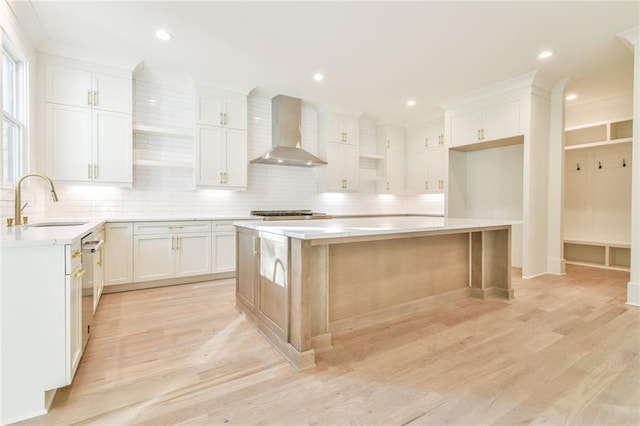 kitchen with wall chimney range hood, sink, white cabinetry, a kitchen island, and light wood-type flooring