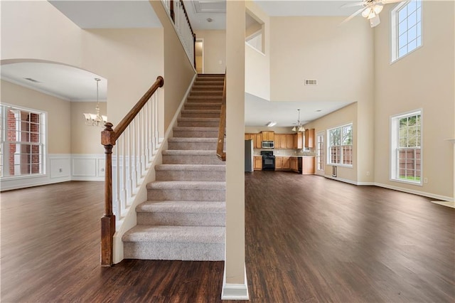 stairs with a wainscoted wall, visible vents, a high ceiling, wood finished floors, and ceiling fan with notable chandelier