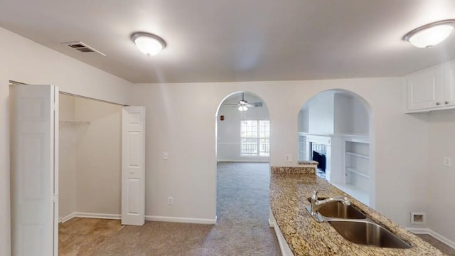 kitchen with arched walkways, a fireplace with raised hearth, a sink, light stone counters, and ceiling fan