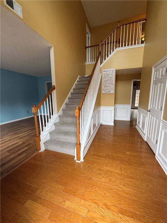 staircase with visible vents, a decorative wall, a towering ceiling, wainscoting, and wood finished floors