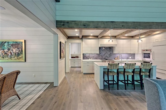 kitchen with a kitchen island with sink, light hardwood / wood-style floors, tasteful backsplash, decorative light fixtures, and beam ceiling