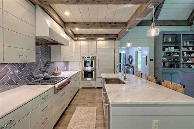 kitchen featuring a center island with sink, appliances with stainless steel finishes, beamed ceiling, decorative light fixtures, and light hardwood / wood-style floors