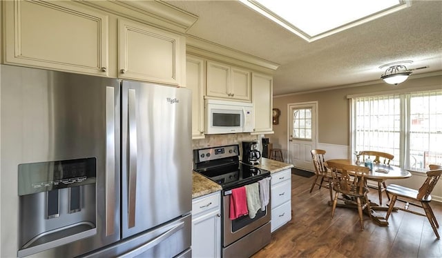 kitchen with a textured ceiling, a wainscoted wall, appliances with stainless steel finishes, cream cabinetry, and dark wood-style flooring