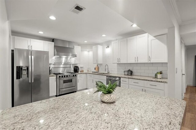 kitchen with wall chimney exhaust hood, hardwood / wood-style floors, light stone counters, appliances with stainless steel finishes, and white cabinetry