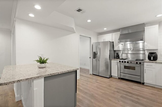 kitchen with light stone countertops, white cabinets, wall chimney exhaust hood, light hardwood / wood-style floors, and appliances with stainless steel finishes