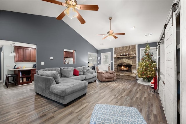 living room with a fireplace, ceiling fan, wood-type flooring, a barn door, and high vaulted ceiling