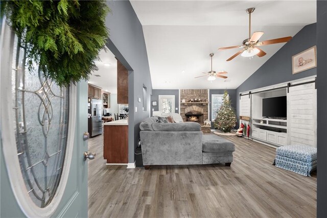 living room featuring a stone fireplace, a barn door, ceiling fan, high vaulted ceiling, and light hardwood / wood-style flooring