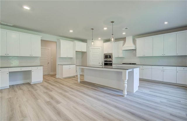 kitchen with premium range hood, white cabinetry, a center island with sink, decorative light fixtures, and light hardwood / wood-style flooring