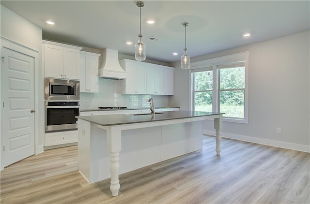 kitchen with decorative light fixtures, premium range hood, a center island with sink, stainless steel appliances, and white cabinets