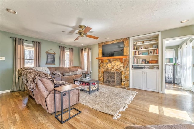 living room with baseboards, light wood-type flooring, a stone fireplace, a textured ceiling, and a ceiling fan