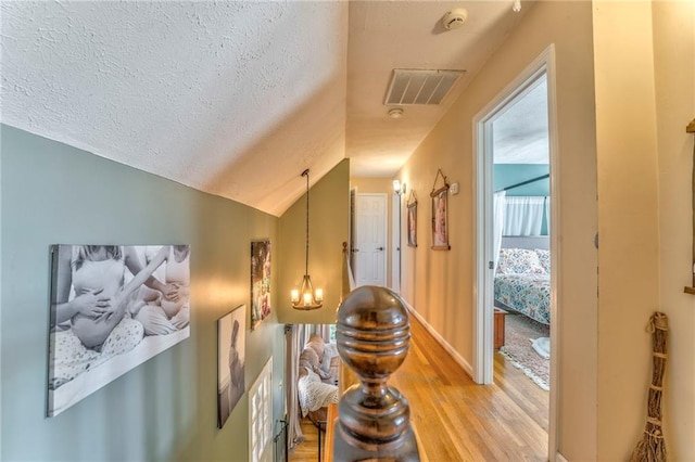 hallway featuring visible vents, vaulted ceiling, light wood-style floors, a notable chandelier, and a textured ceiling