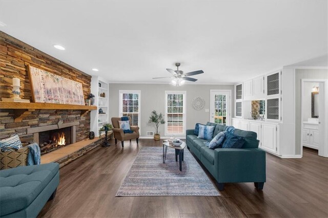 living area featuring built in shelves, crown molding, ceiling fan, a stone fireplace, and dark wood-style flooring