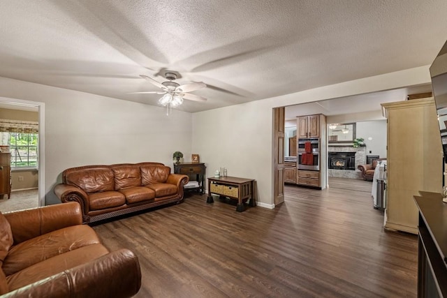 living room featuring a ceiling fan, a warm lit fireplace, a textured ceiling, baseboards, and dark wood-type flooring