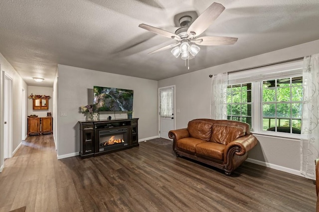 living area with a textured ceiling, dark wood-style flooring, baseboards, ceiling fan, and a glass covered fireplace