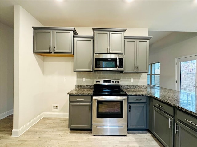 kitchen with decorative backsplash, gray cabinets, dark stone countertops, light wood-type flooring, and stainless steel appliances