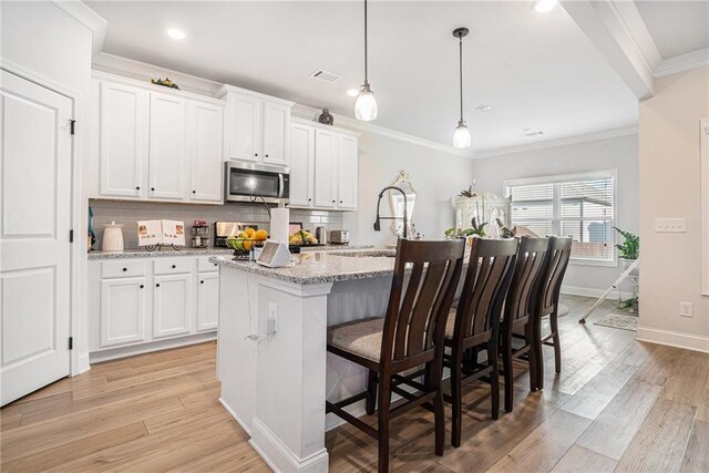 kitchen featuring decorative backsplash, an island with sink, white cabinetry, stainless steel microwave, and light wood finished floors
