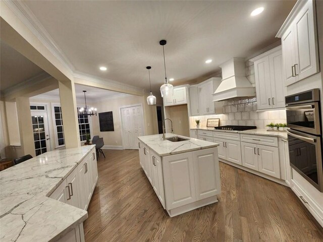 kitchen featuring sink, custom exhaust hood, light stone counters, a center island with sink, and white cabinets