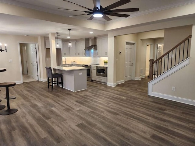 kitchen featuring dark wood-type flooring, wall chimney exhaust hood, decorative light fixtures, a center island with sink, and stainless steel appliances