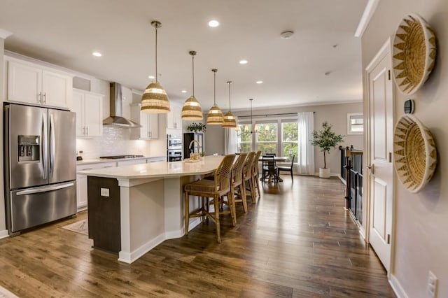 kitchen with stainless steel appliances, a kitchen island with sink, wall chimney range hood, white cabinetry, and hanging light fixtures