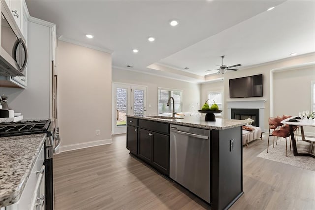 kitchen featuring a kitchen island with sink, stainless steel appliances, a sink, light wood-type flooring, and a tray ceiling