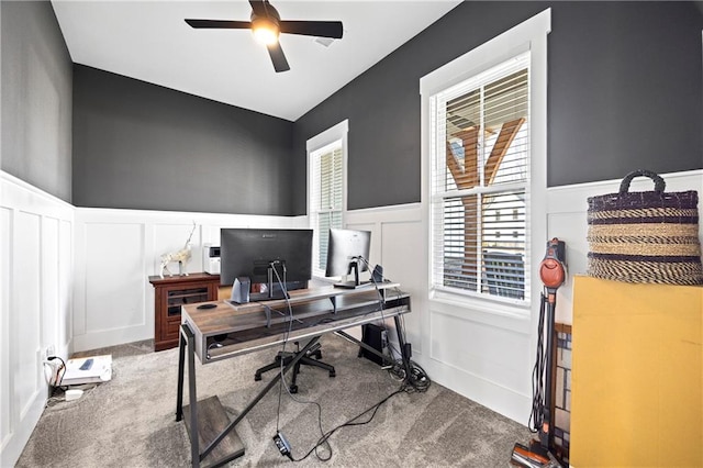 carpeted home office with a wainscoted wall, ceiling fan, and a decorative wall